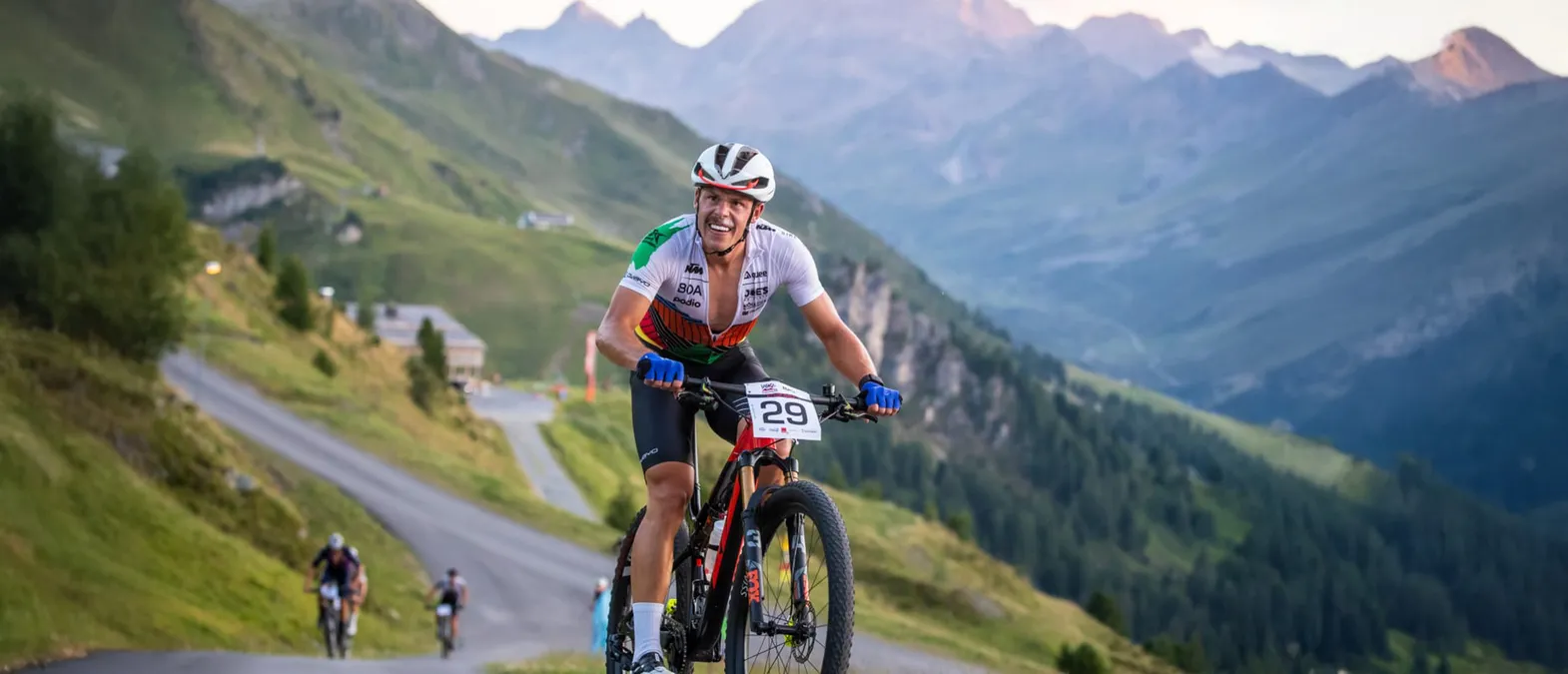 Een fietser in een wit, groen en oranje tricot met nummer 29 rijdt op een bergweg in een bergachtig landschap.