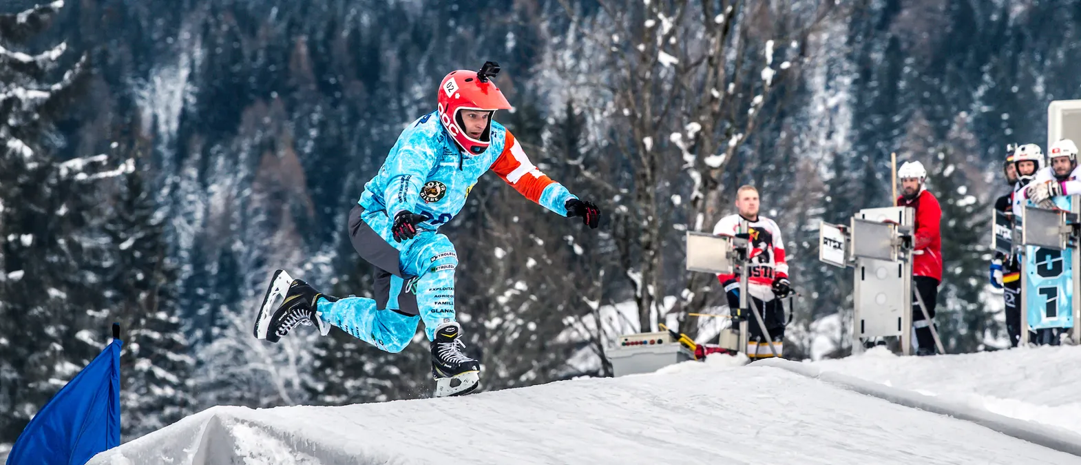 Een ijshockeyspeler in een blauw-wit, met sponsorlogo's bedrukt shirt en een rode helm springt over een sneeuwheuvel op een ijsbaan, met andere spelers en bomen op de achtergrond.