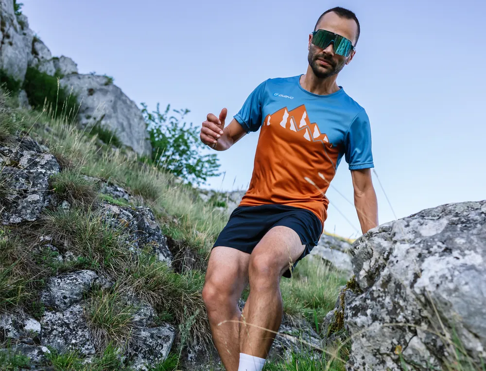 Een man rent over een rotsachtig pad in een sportief blauw-oranje T-shirt en shorts.