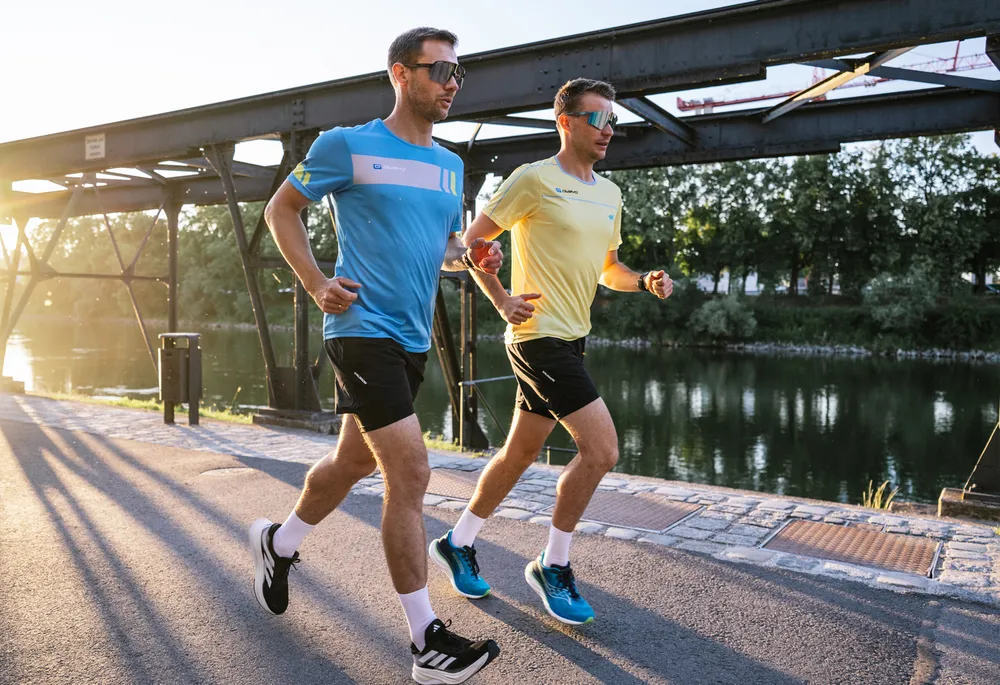 Twee mannen in sportkleding joggen langs een kalme waterpartij terwijl de zon opkomt.