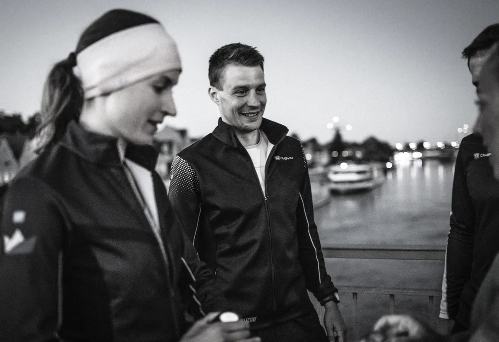 Twee personen dragen sportjacks en kletsen ontspannen bij een waterlichaam tijdens de schemering.