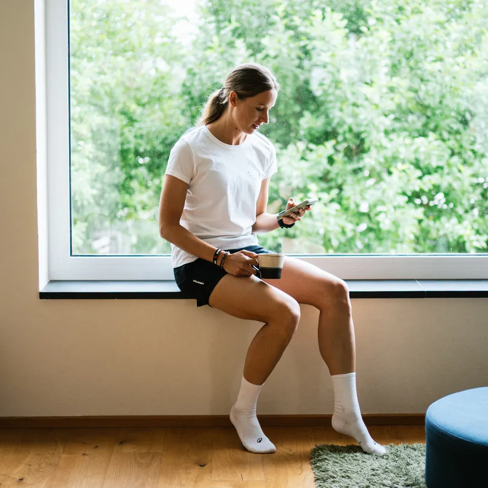 Een vrouw zit op een vensterbank, gekleed in een wit T-shirt en zwarte shorts, terwijl ze een smartphone vasthoudt en geniet van een kop koffie.