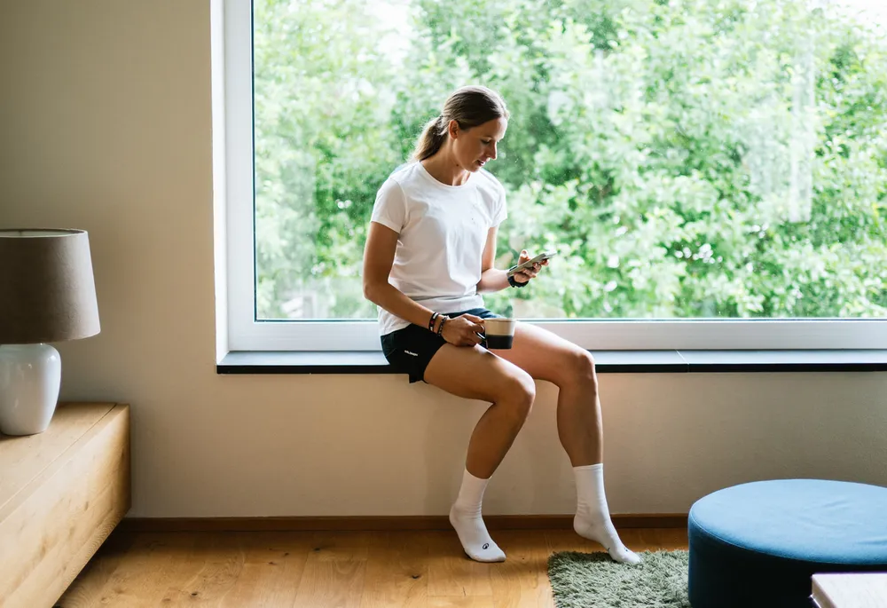 Een vrouw zit op een vensterbank met een telefoon in de ene hand en een kopje in de andere, gekleed in een wit shirt en zwarte shorts.