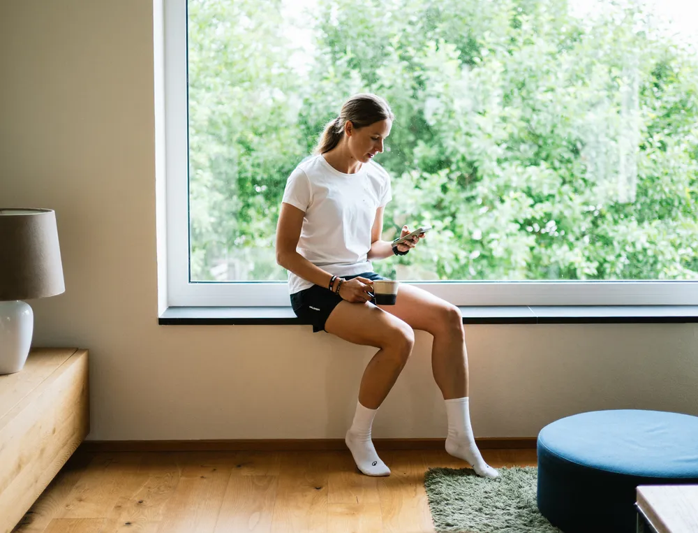 Een vrouw zit op een vensterbank, kijkt op haar telefoon terwijl ze een kop koffie vasthoudt, gekleed in een wit T-shirt en zwarte shorts.