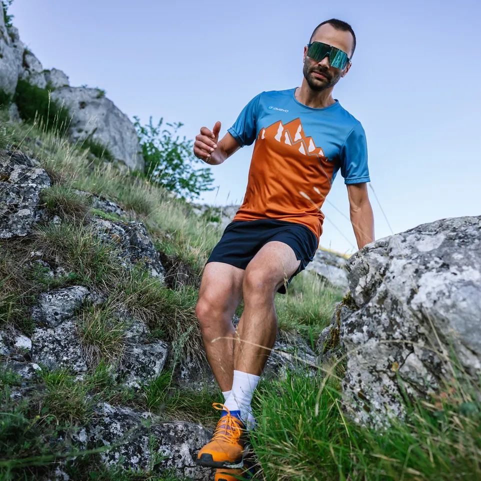 Een man rent op een rotsachtig pad en draagt een blauw-oranje sportshirt met korte mouwen met een bergontwerp en een zwarte short.