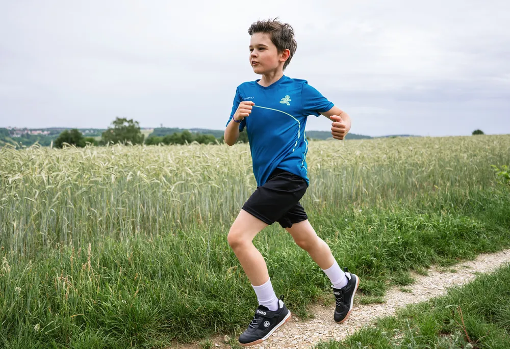 Een jongen in een blauw sportshirt en zwarte shorts rent langs een pad door een veld.