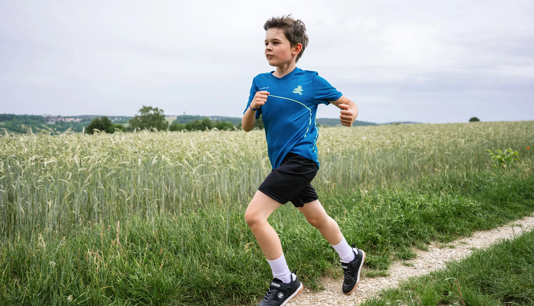 Een jongen rent langs een pad door een veld, gekleed in een blauw sportshirt en zwarte shorts.