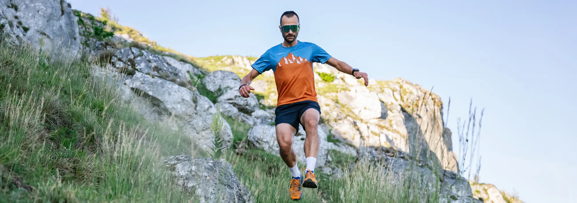 Een mannelijke hardloper in een oranje-blauw shirt en shorts, die een rotsachtige helling afloopt.