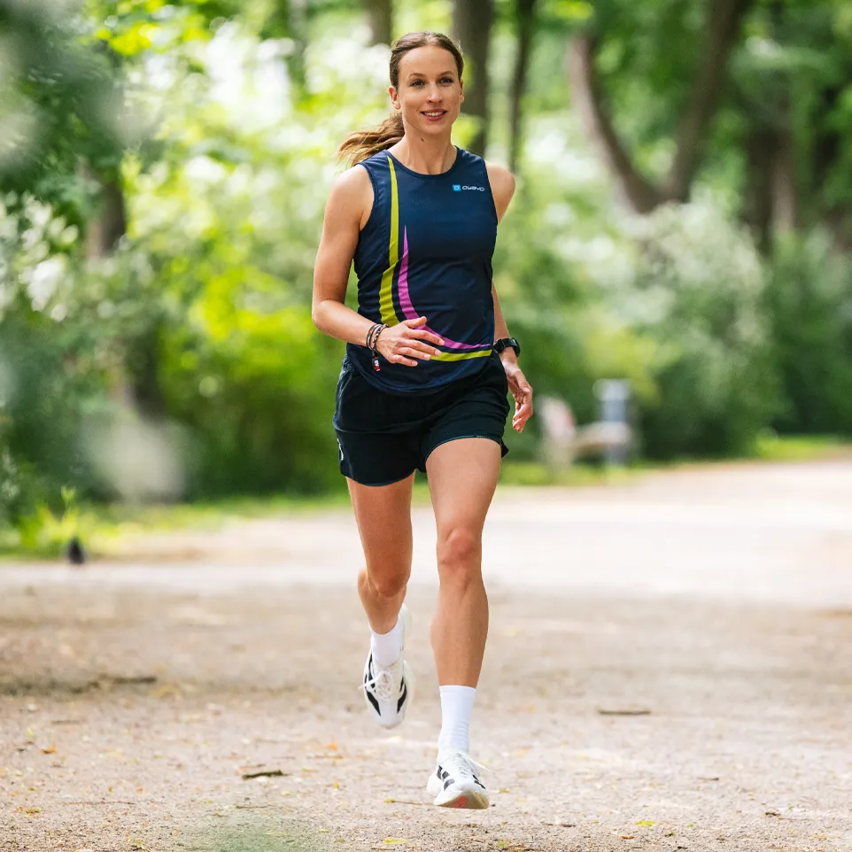 Een sportieve vrouw loopt op een pad in een park, gekleed in een mouwloos donkerblauw shirt met kleurrijke accenten en shorts.