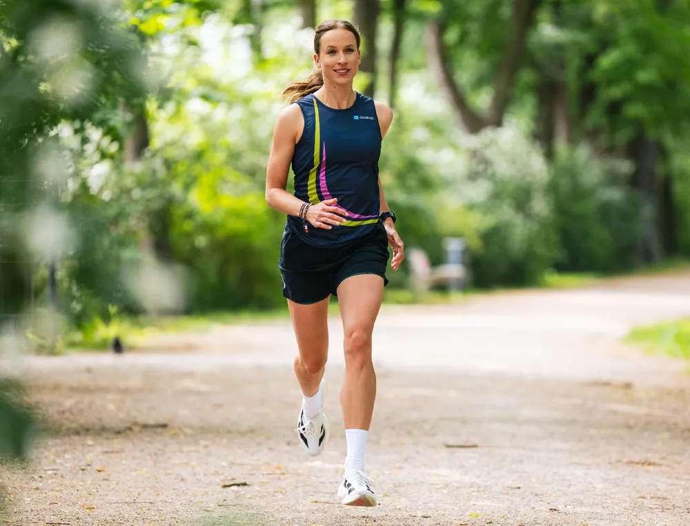 Een sportieve vrouw rent langs een smal pad door een groen park, gekleed in een blauwe tanktop met kleurrijke accenten en shorts.