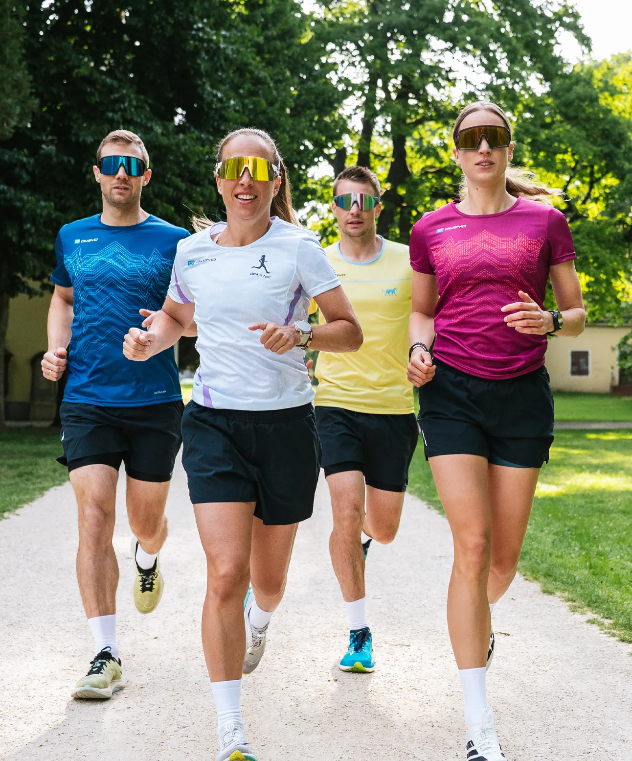 Vier atletische personen rennen op een smal pad in een park, gekleed in kleurrijke hardloopshirts en zonnebrillen.