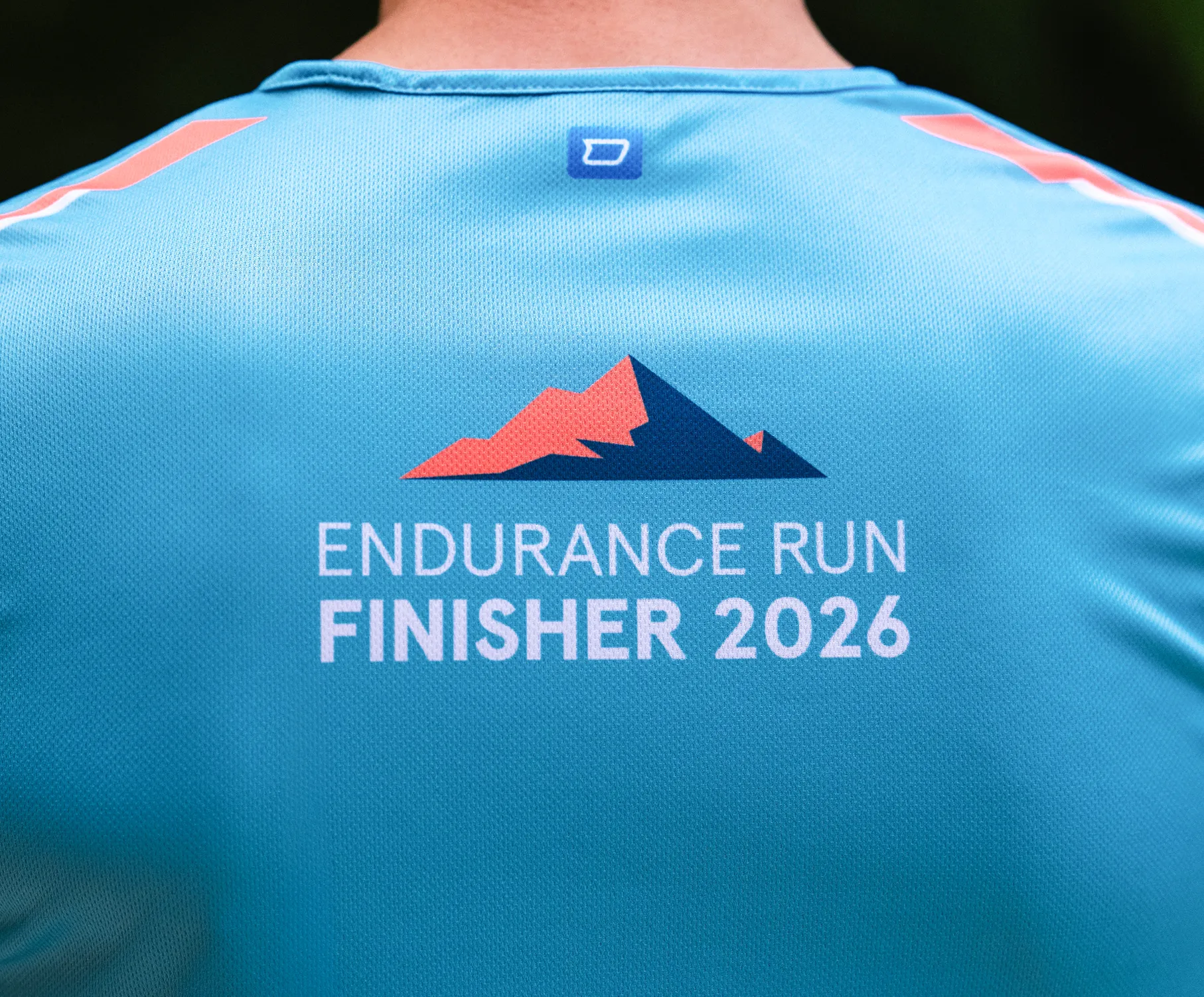 Een blauw sportshirt met de opdruk 'ENDURANCE RUN FINISHER 2026' en een gestileerd bergontwerp op de achterkant.
