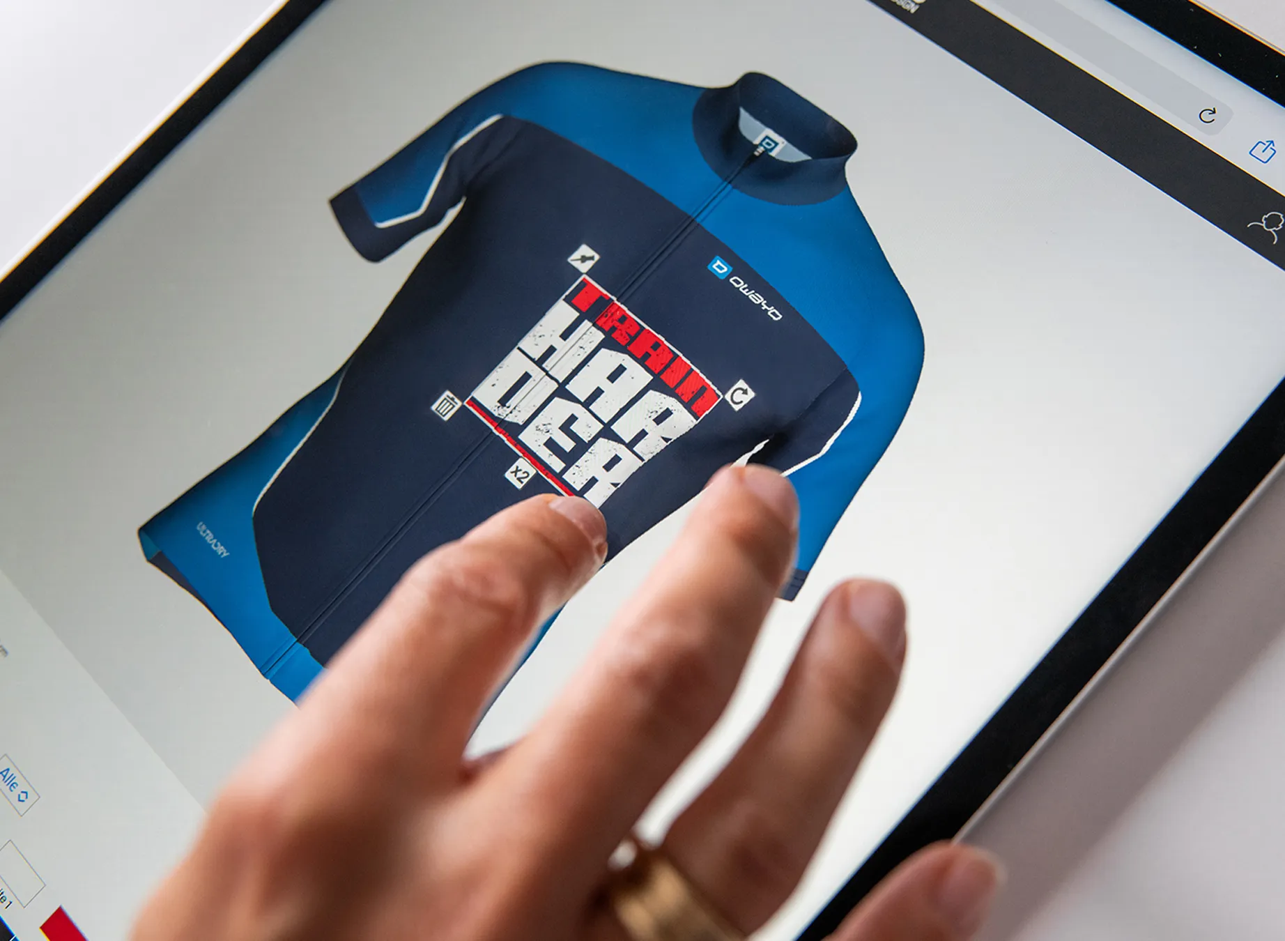 Een hand wijst naar een digitaal ontwerp van een blauwe sporttrui met de tekst 'train harder'.
