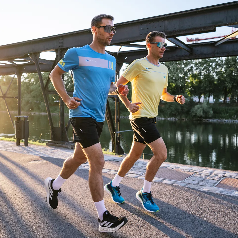 Twee mannen joggen op een pad langs een waterlichaam, de een draagt een blauw T-shirt en de ander een geel T-shirt.