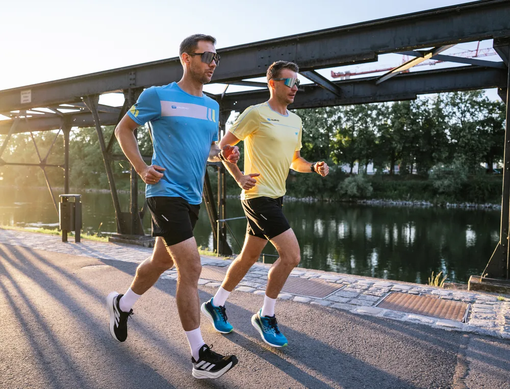 Twee mannen joggen aan de waterkant, de een draagt een blauwe t-shirt en de ander een gele, beiden in korte broeken.