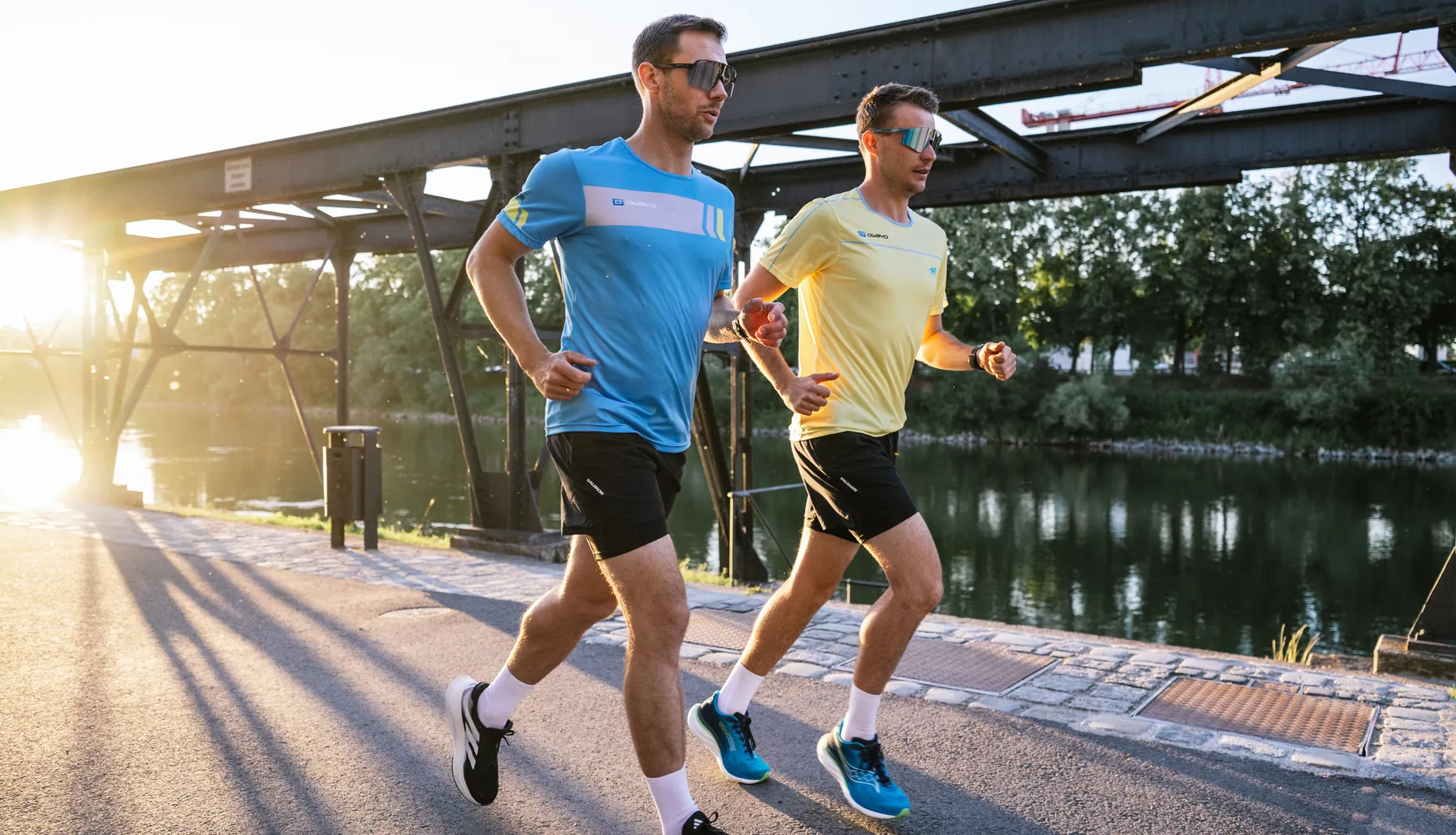 Twee mannen in sportieve hardloopkleding, de een in een blauwe t-shirt en de ander in een gele t-shirt, joggen langs de oever van een waterweg bij zonsondergang.