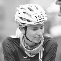 Een vrouwelijke fietser in een zwart fietsshirt met het logo 'IWC Schaffhausen', een witte helm met nummer 183 en een sjaal om haar nek.