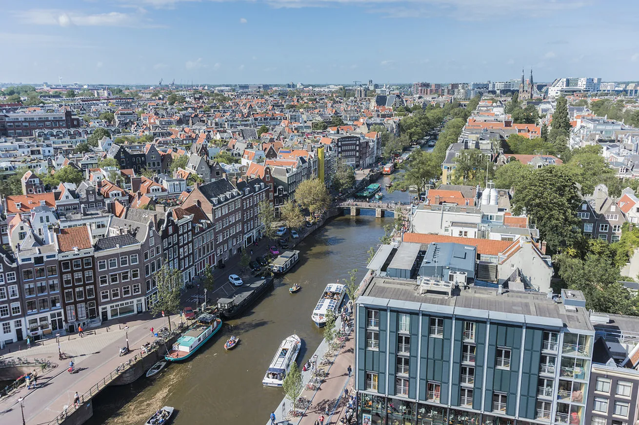 Luchtfoto van Amsterdam met grachten, traditionele gebouwen en boten onder een blauwe lucht.
