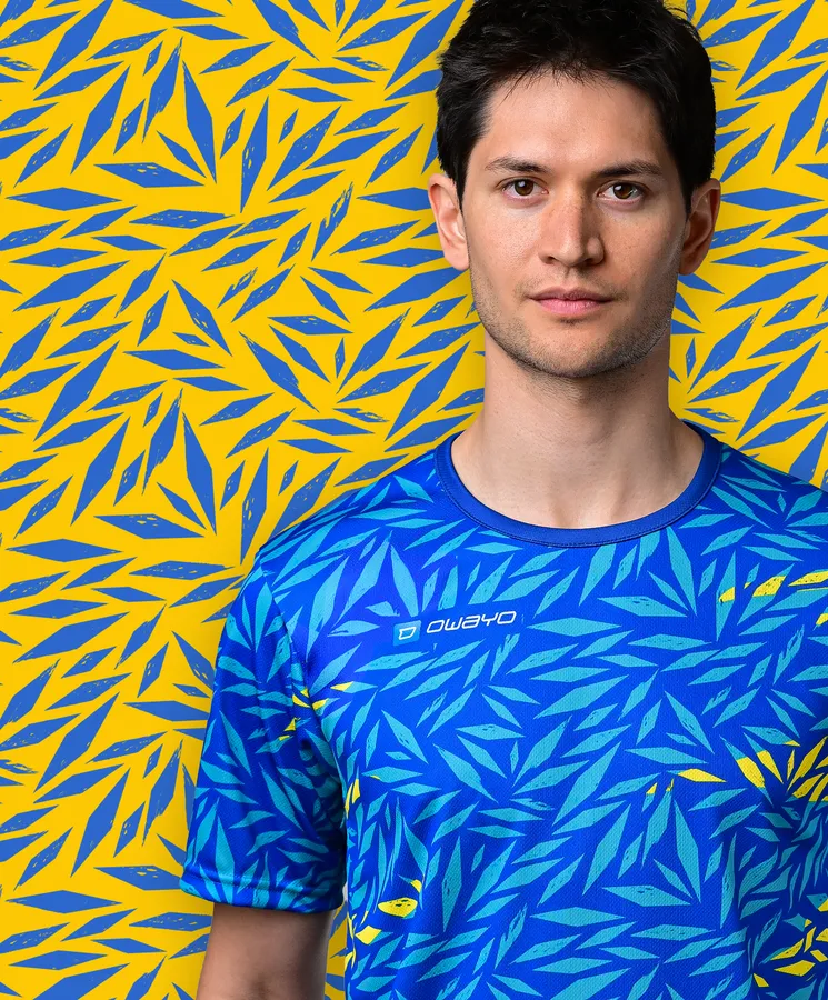 Een jonge man draagt een sportief blauw T-shirt met een kleurrijk bladmotief tegen een gele achtergrond.