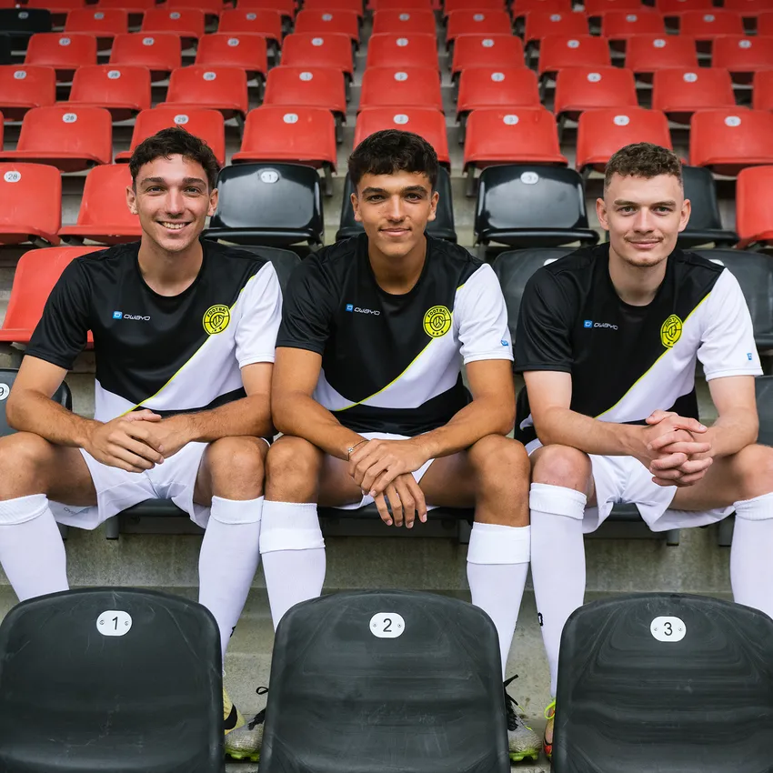 Drie jonge mannen in owayo sportkleding zitten in een stadion op genummerde stoelen voor rode tribunes.