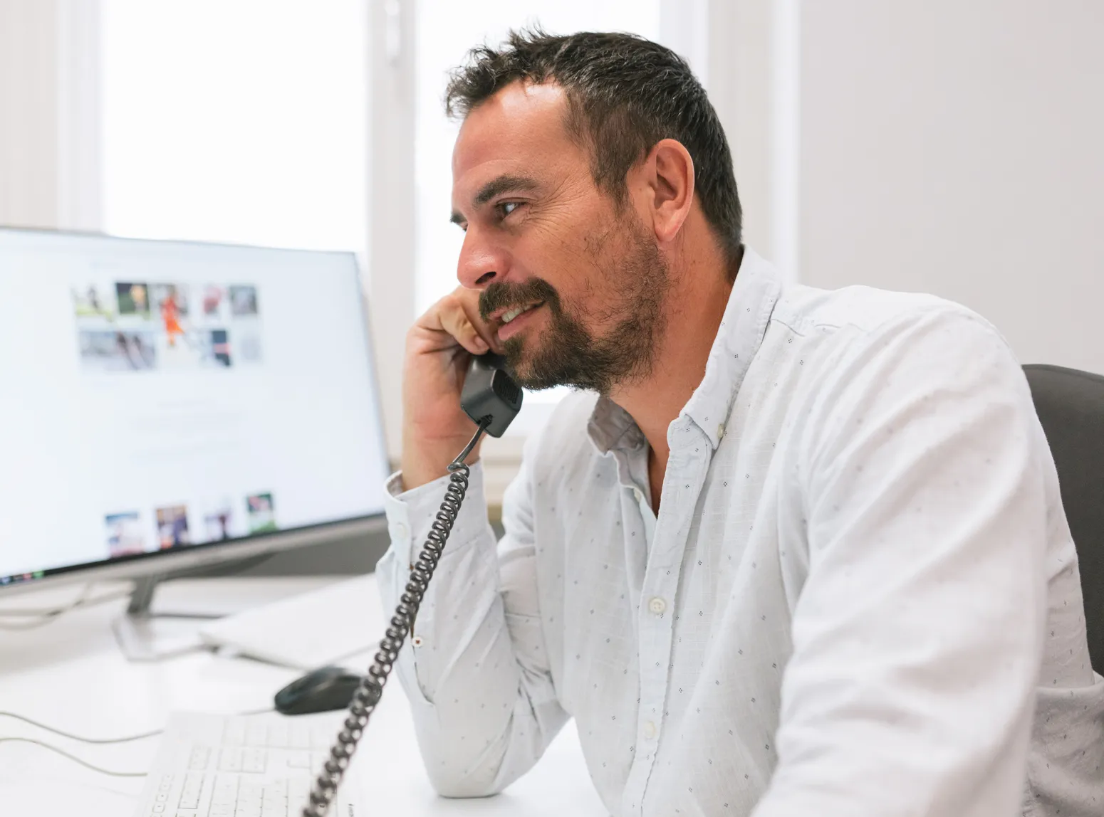 Een man met een baard zit aan een bureau voor een computer en glimlacht terwijl hij telefoneert met een ouderwetse telefoonhoorn.