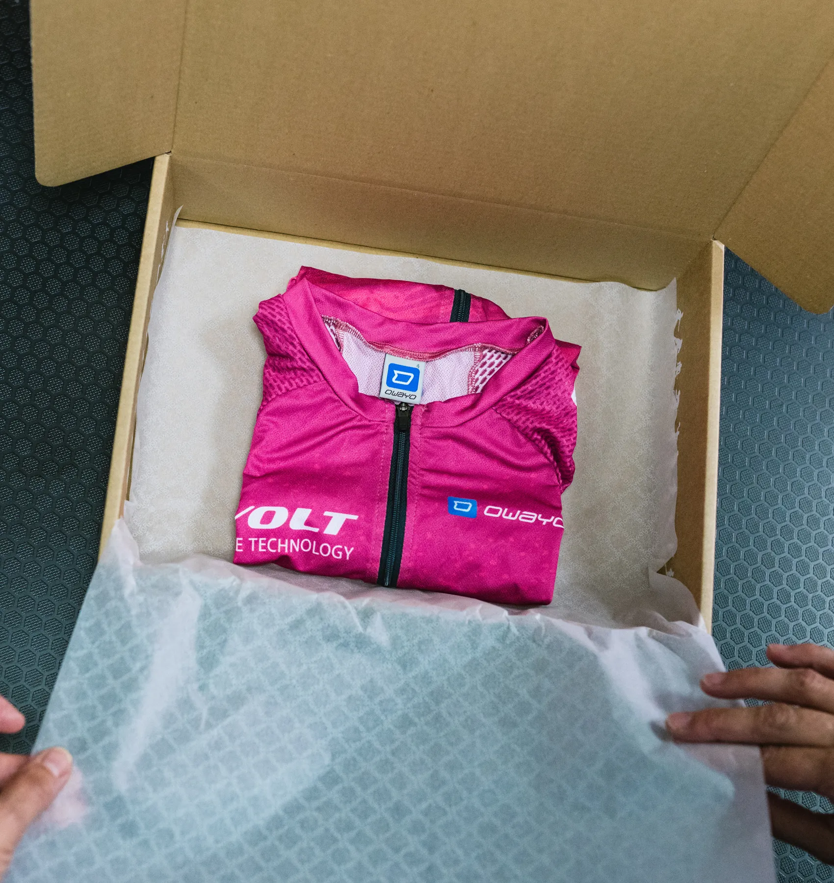 Een opgevouwen, roze, op maat gemaakte sportkleding van owayo ligt in een open kartonnen doos op wit papier, vastgehouden door handen.
