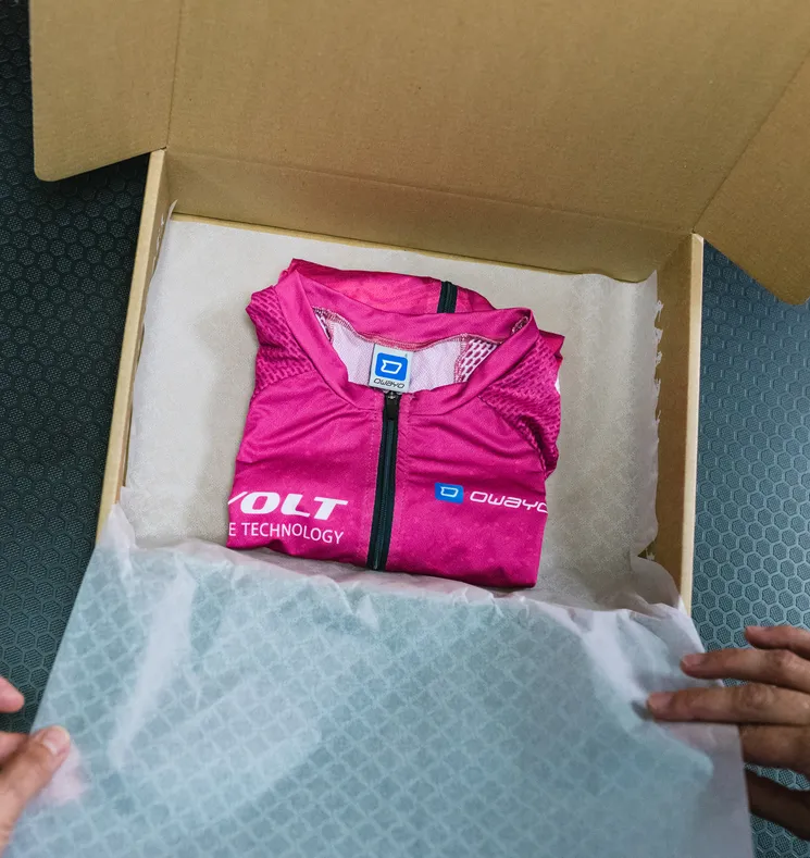 Een opgevouwen, roze, op maat gemaakte sportkleding van owayo ligt in een open kartonnen doos op wit papier, vastgehouden door handen.