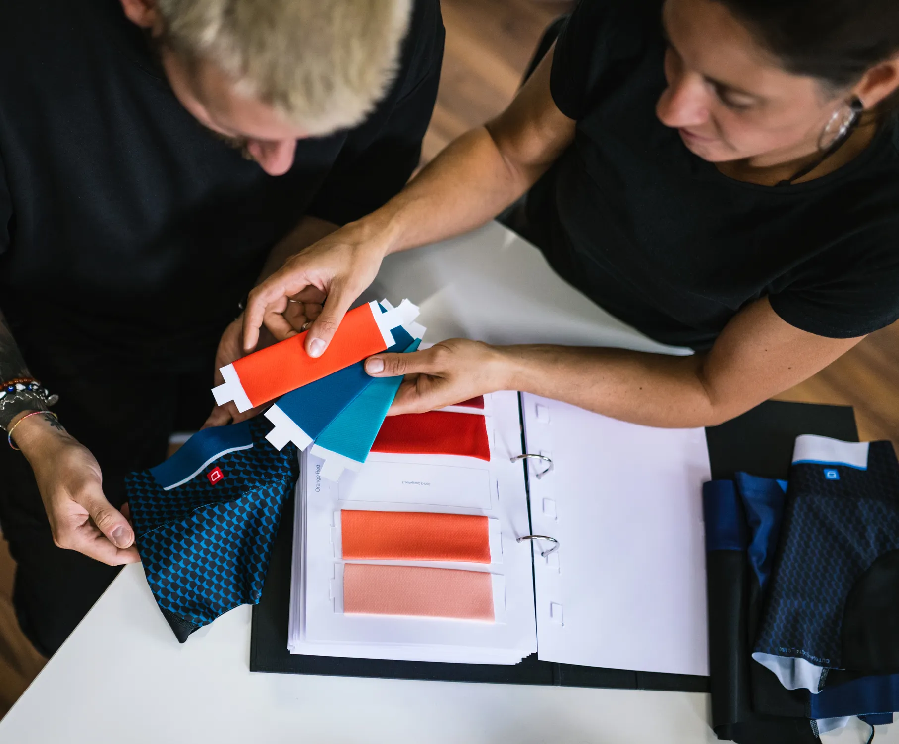 Twee personen kiezen kleuren voor aangepaste sportkleding door stofmonsters en een kleurenschakering te bekijken.