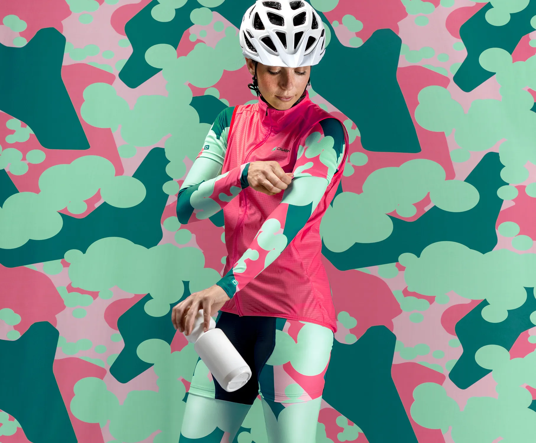 Een fietser in een aangepaste roze-groene sportkleding met een abstract patroon, die haar mouwen aanpast terwijl ze een witte fles vasthoudt, tegen een kleurrijke achtergrond met hetzelfde patroon.