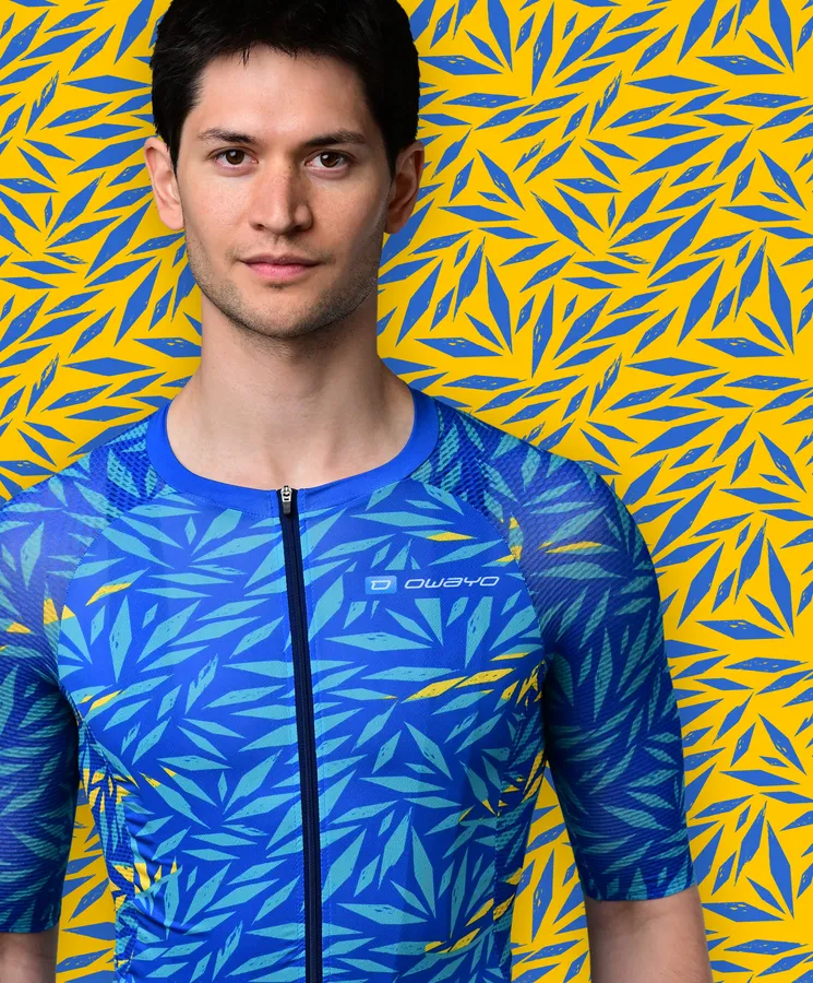 Een man draagt een blauw, korte mouwen sportshirt met een blauw en geel bladpatroon dat doet denken aan een tropische plant, tegen een gele achtergrond met blauwe bladpatronen.