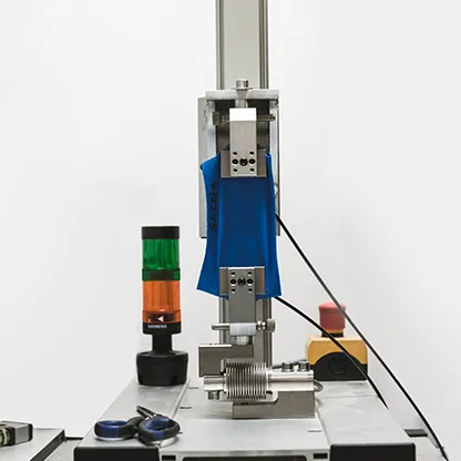 Een blauwe stof wordt getest in een machine op een werkbank, naast een signaallamp en gereedschap.