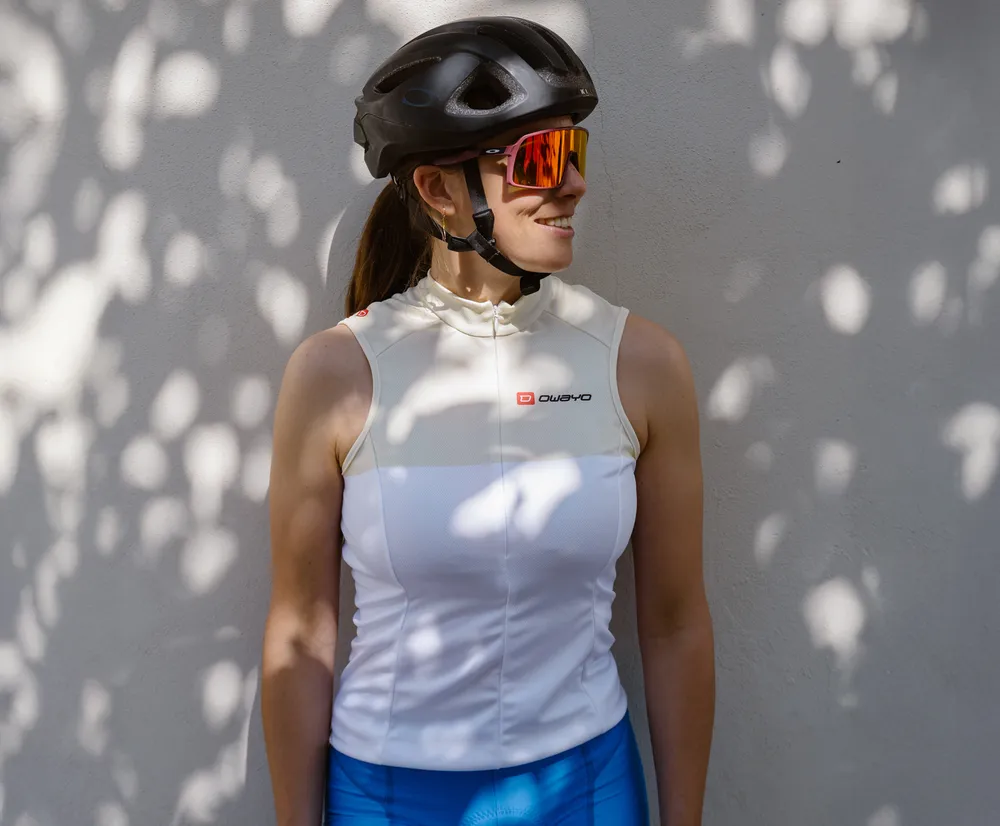 Een vrouw in een wit, mouwloos fietsshirt met het owayo-logo, een fietshelm en zonnebril, leunend tegen een muur met bladschaduwen.