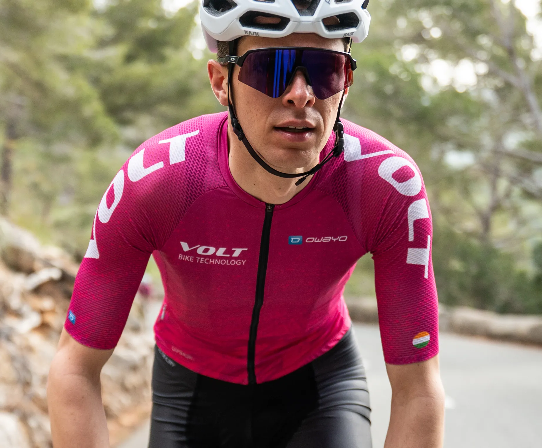 Een fietser in een roze, korte mouwen fietsshirt met het logo 'VOLT BIKE TECHNOLOGY' en het 'owayo'-logo op de borst, die op een weg in een bosrijke omgeving fietst.