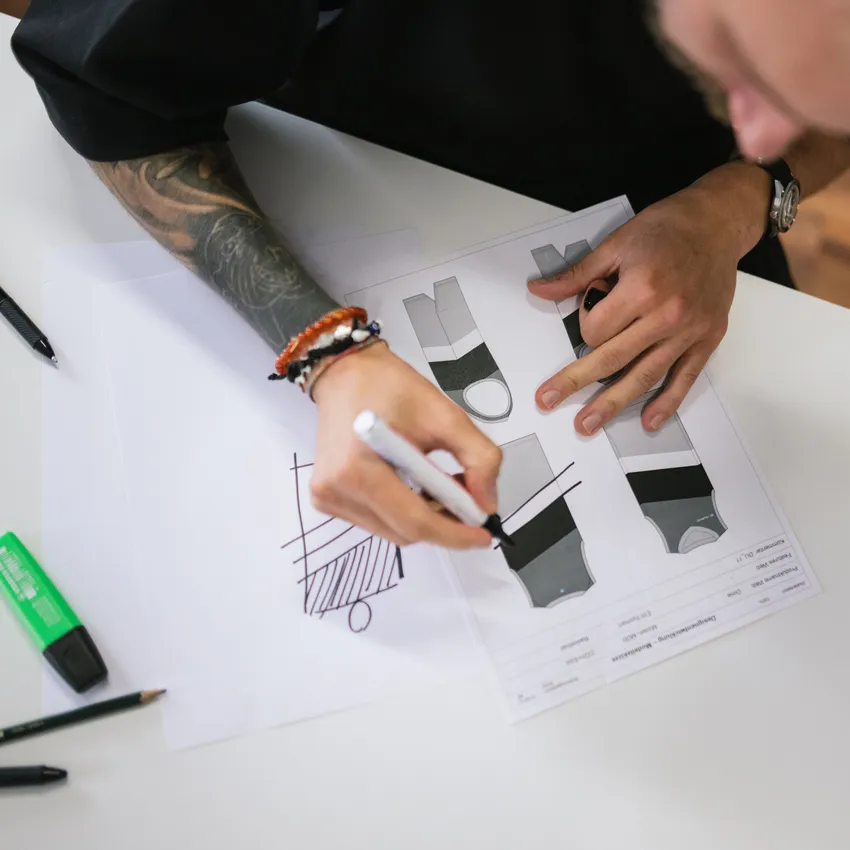 Een persoon maakt schetsen van ontwerpen voor sportkleding op papier, omringd door pennen en markeerstiften.