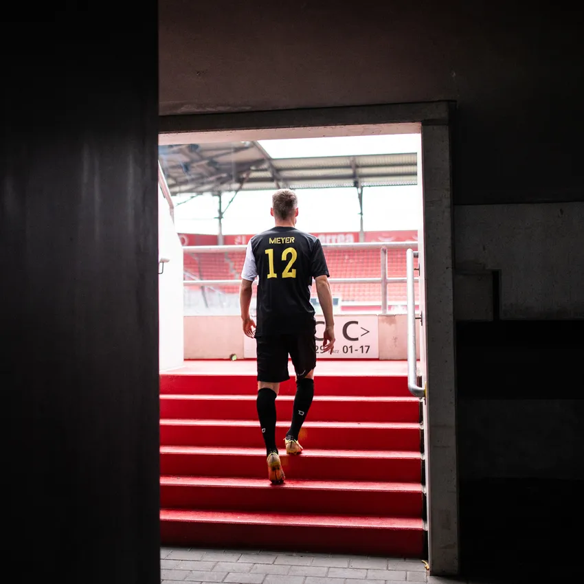 Een voetballer in een zwart shirt met het nummer 12 loopt de rode trappen van een stadion op.