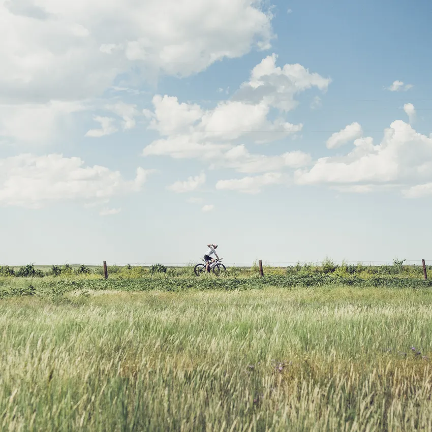 Een persoon fietst door een groene landschap onder een blauwe lucht met enkele wolken.