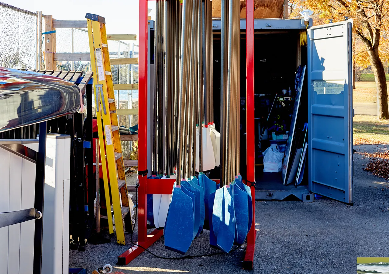Een afbeelding van een botenopslagplaats met roeiriemen in verschillende kleuren, een gele ladder en een open container op de achtergrond.
