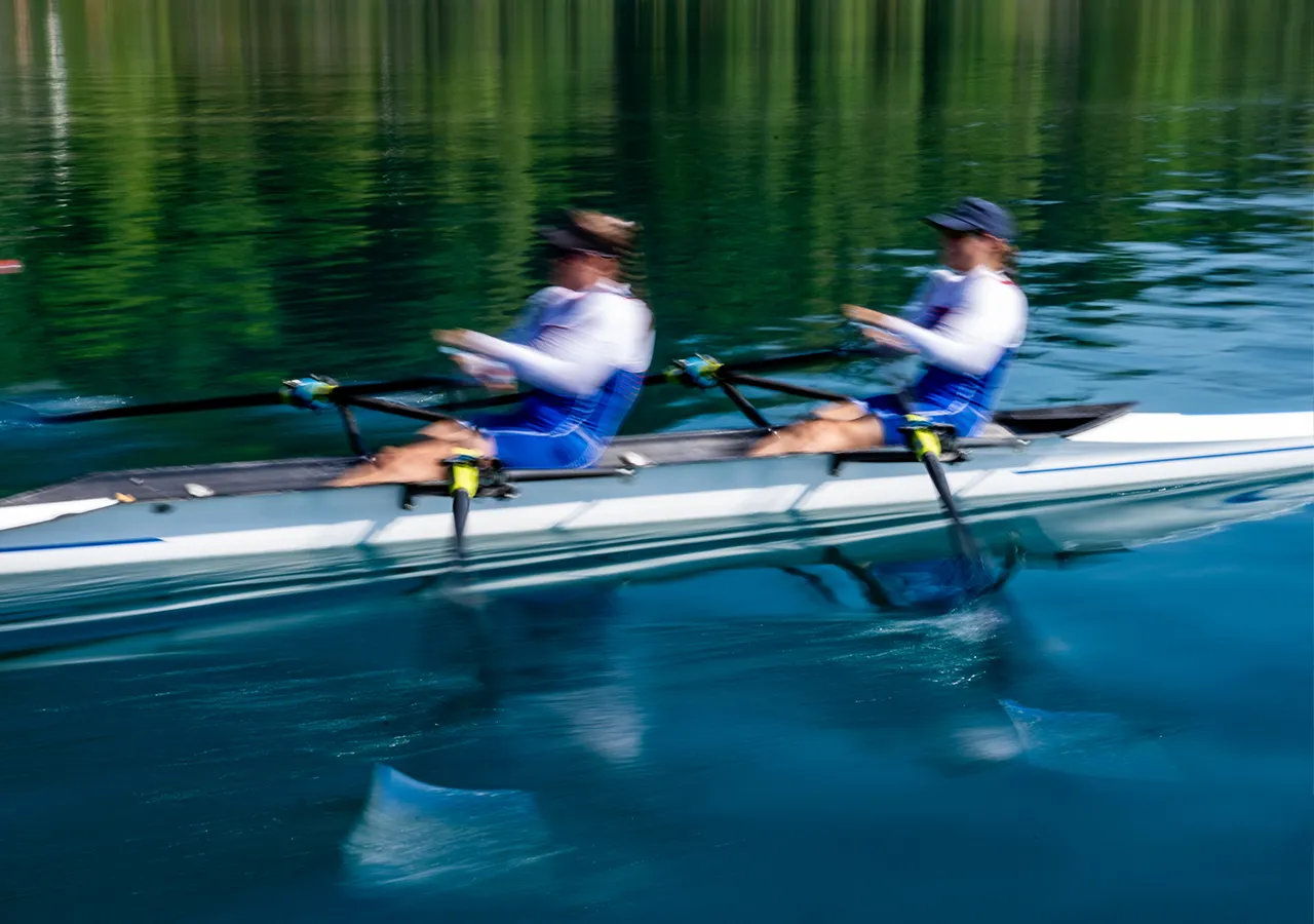 Twee roeiers in blauwe sportkleding roeien synchroon op een kalme wateroppervlakte.