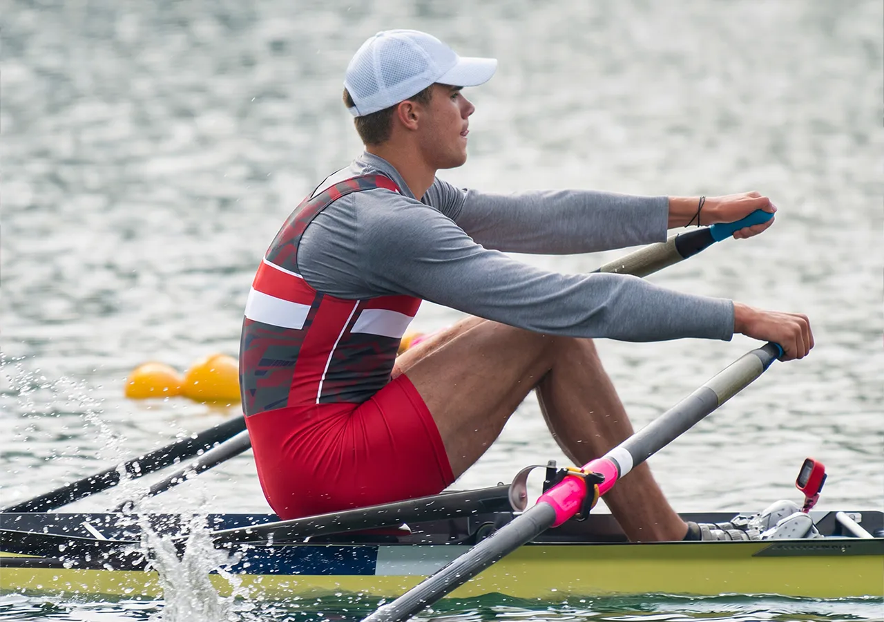 Een roeier in een rood-grijze sportoutfit roeit op een wateroppervlak.