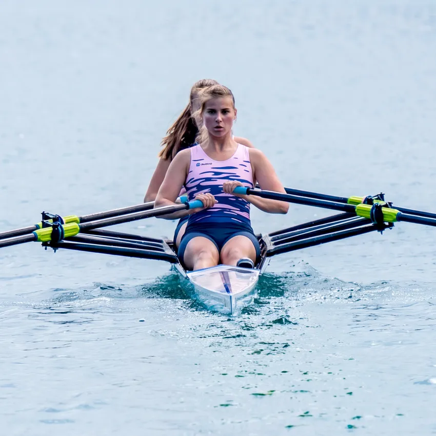 Twee roeiers in een boot die op kalm water roeien, waarbij de voorste roeier een roze sportoutfit draagt.
