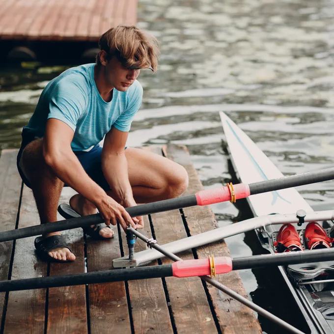 Een jonge man in een blauw sportshirt repareert roeispanen op een dock aan het water.