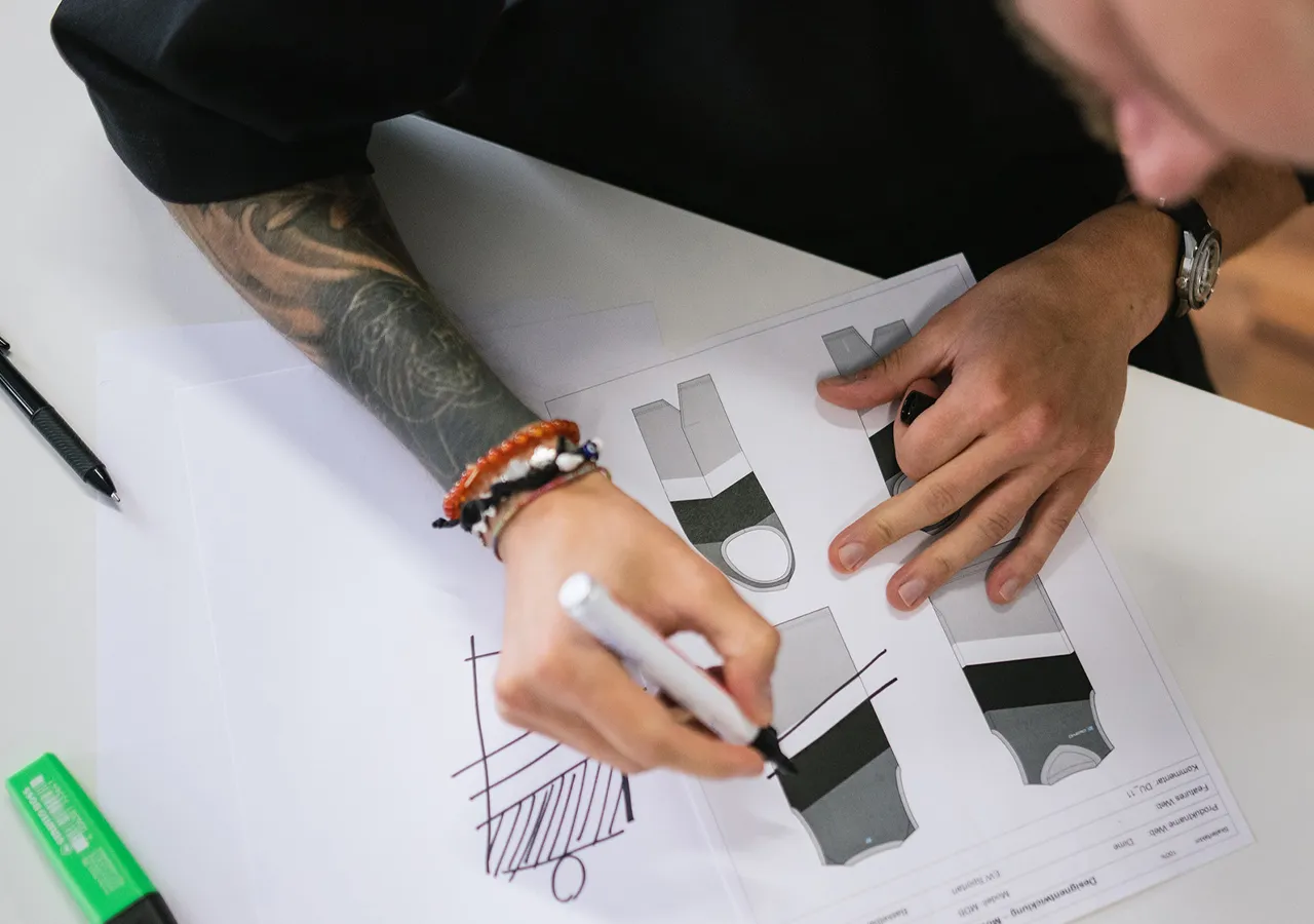 Een persoon met een tatoeage op de arm tekent met een witte marker op een vel papier met meerdere ontwerpen van sportkleding.