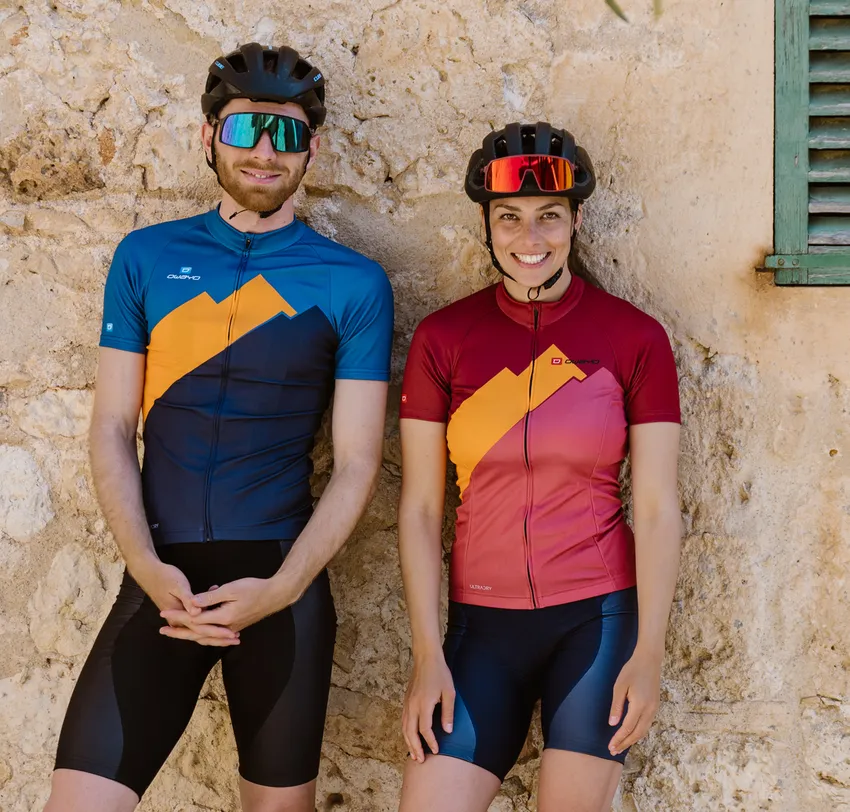 Een man en een vrouw in aangepaste fietskleding van owayo, staand voor een stenen muur.