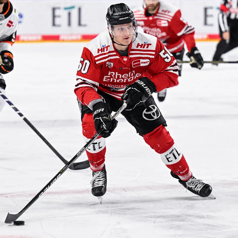 Een ijshockeyspeler in een rood shirt met nummer 50 en de tekst 'Rhein Energie' op het ijs, die een puck beheert met zijn stok.