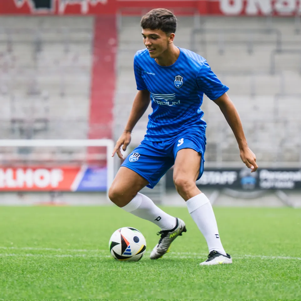 Een jonge voetballer in een blauw shirt en blauwe broek met een logo op de borst en het nummer 3 op de broek dribbelt een voetbal op een groen grasveld.