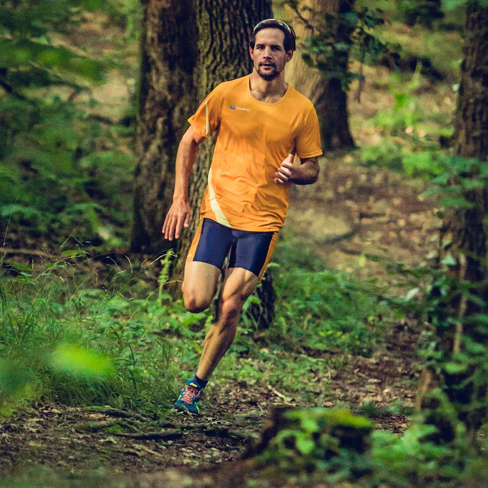 Een man loopt op een bospad in een oranje hardloopshirt en donkerblauwe hardloopshorts.