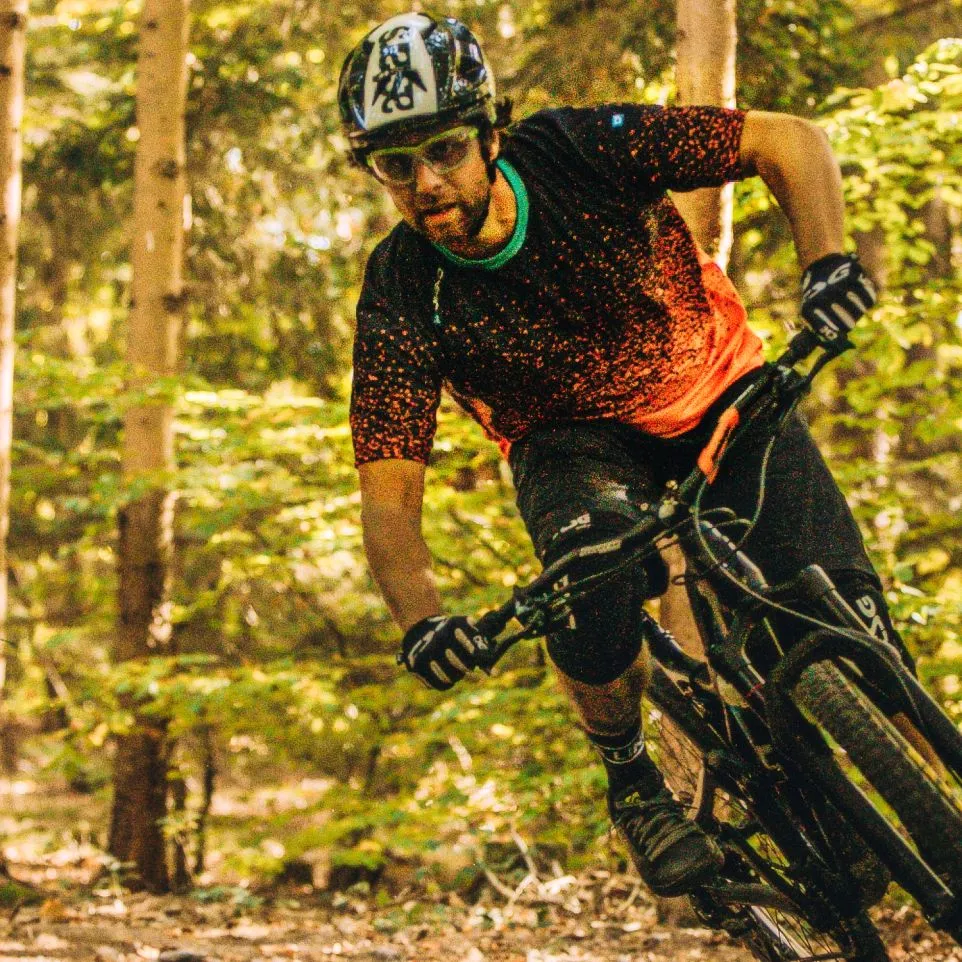 Een mountainbiker in een zwart, rood gestippeld shirt en zwarte broek fietst door een bos.