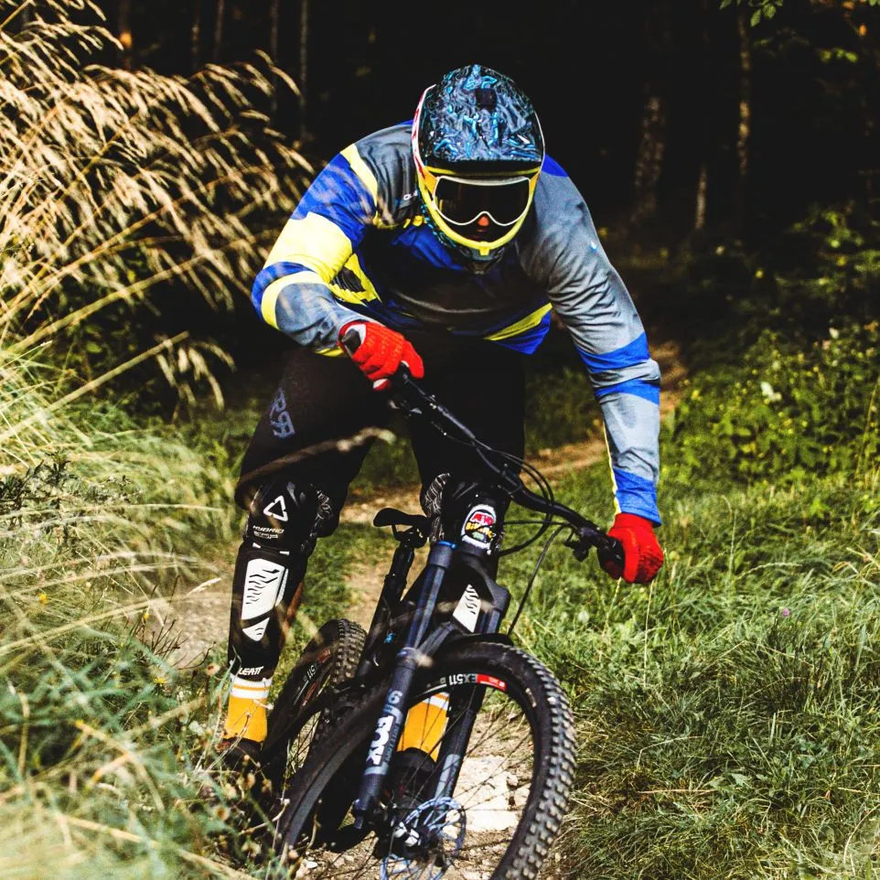 Een mountainbiker in een kleurrijk, lang mouwtrikot met blauwe, gele en grijze accenten rijdt op een bospad.
