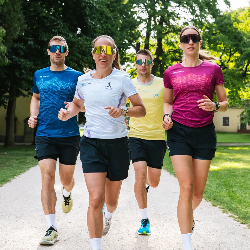 Vier hardlopers in kleurrijke sportshirts en shorts joggen op een pad in een park.