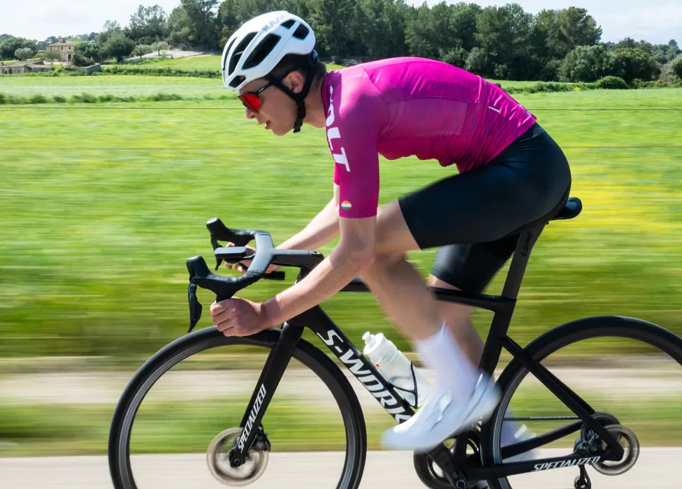 Een fietser in een roze trui en zwarte korte broek fietst op een racefiets op een landweg, met een wazig groen achtergrond dat snelheid suggereert.